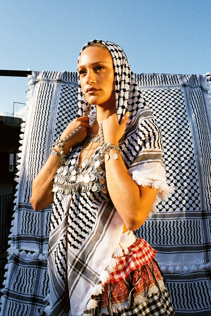 Woman wearing a patterned headscarf and dress against a blue sky with clouds.